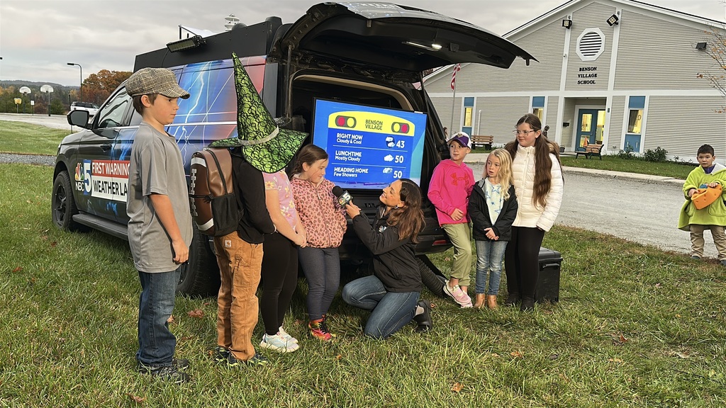 NBC 5 meteorologist kneels beside the weather lab vehicle interviewing children, one wearing a green witch hat, with the Benson Village School building in the background.