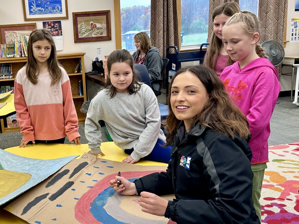 NBC 5 meteorologist sits with students in a school library working on a large painted weather-themed poster together.