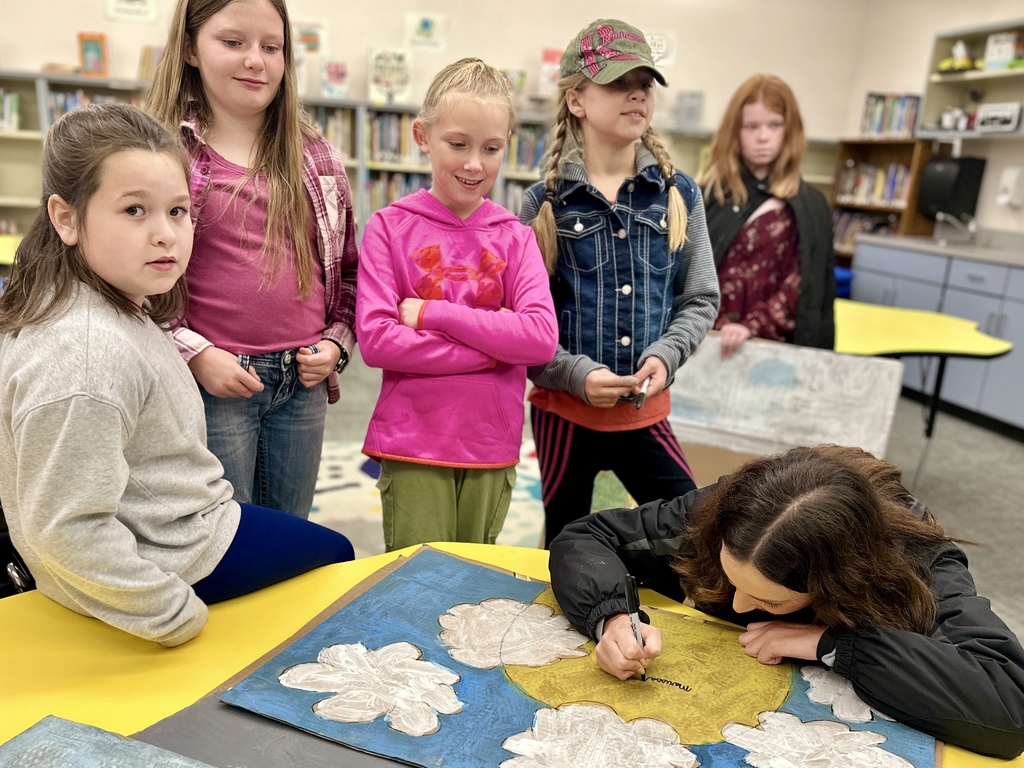 Meteorologist signs a painted cardboard sun and cloud project while several students stand around watching in the library.