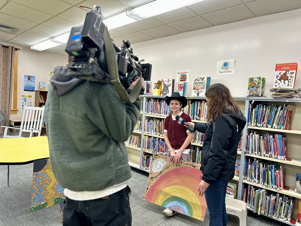 A student being interviewed by the NBC 5 meteorologist in the library holds a painted rainbow project as a cameraman films.