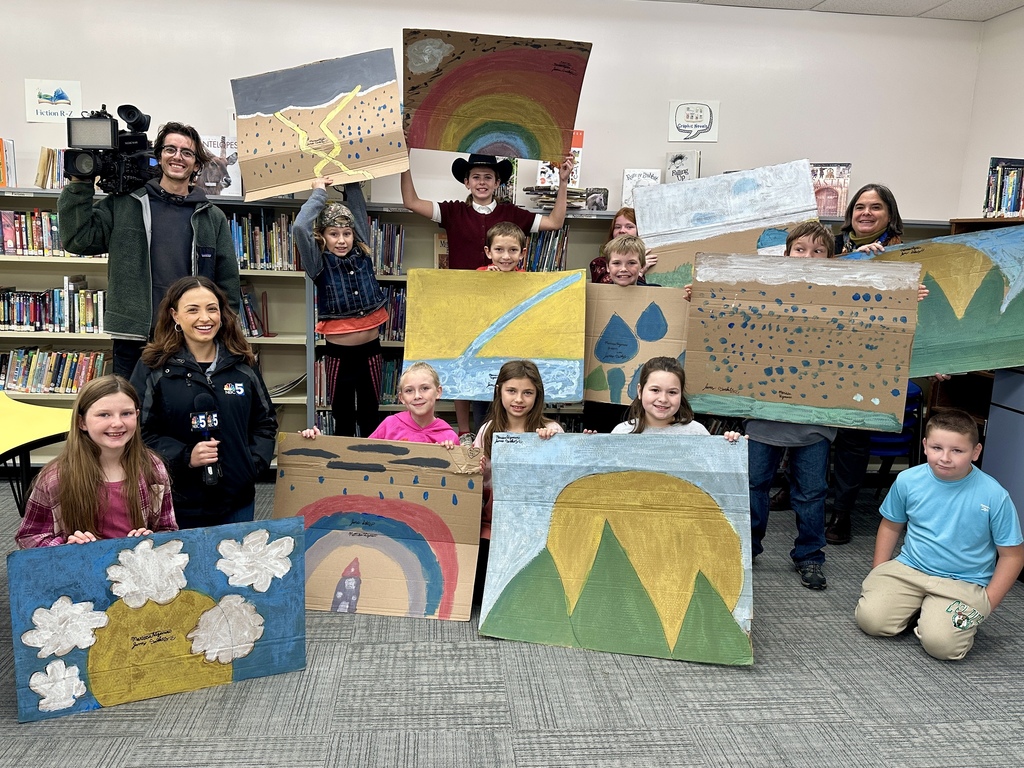 Group of students, teachers, and NBC 5 crew pose in the library holding up colorful weather-themed artwork made on cardboard.