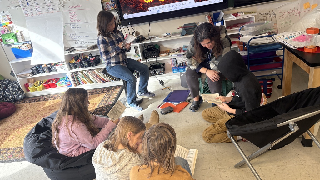 Small group of students and a teacher sit in a circle reading books together on the classroom floor during a group reading activity.