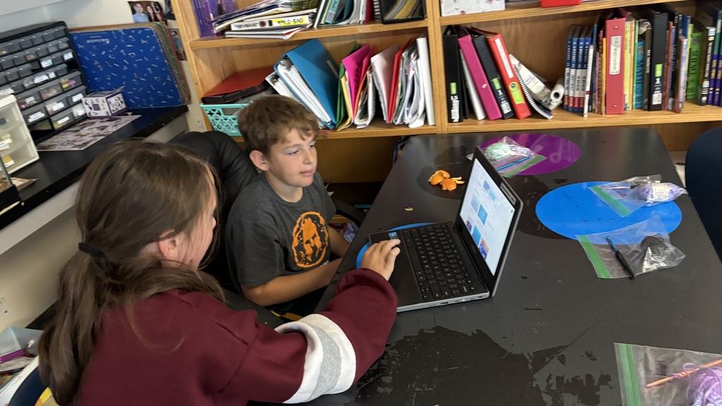 Two students collaborate at a table, looking at a laptop screen together in a classroom with shelves of books and binders.