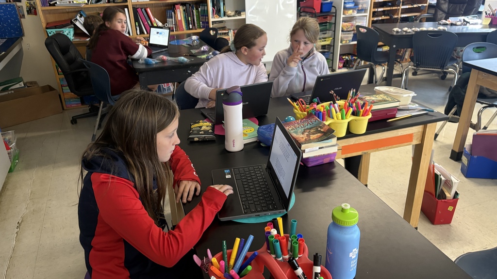Students sit at classroom tables working on laptops surrounded by colorful pens, books, and supplies.