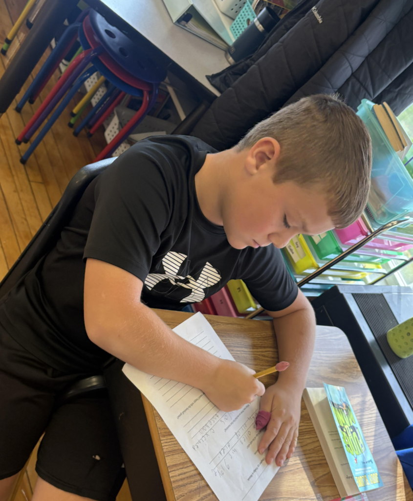 Student wearing a black shirt writes answers on a worksheet at his desk, with colorful bins and school supplies in the background.