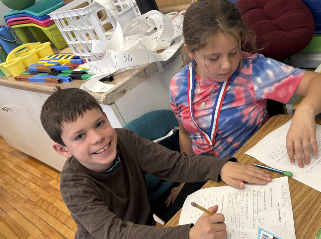 Two students work together on a writing assignment at a classroom table; one smiles at the camera while the other focuses on her paper.