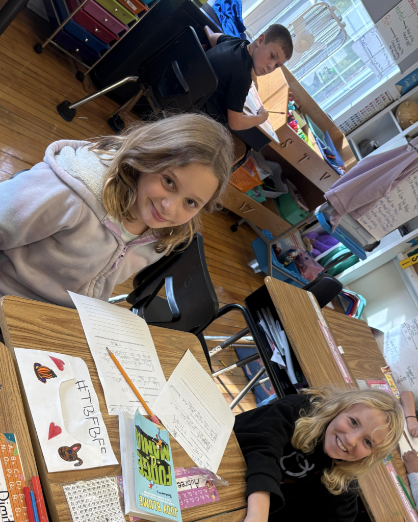 Two smiling students sit at a shared desk with writing papers and a book titled Fudge-a-Mania in front of them.