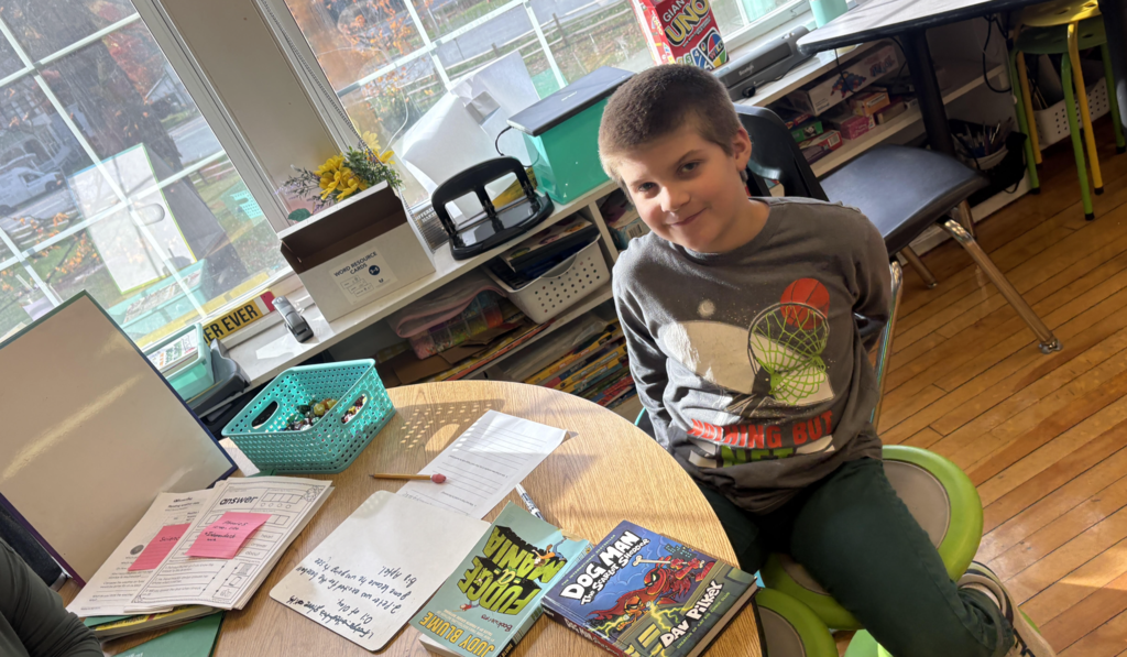 Student sits at a small round table near a window, smiling beside books including Dog Man and Fudge-a-Mania and completed worksheets.