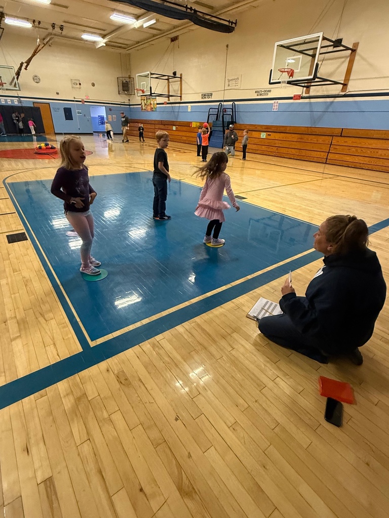 An adult kneels on a wooden gymnasium floor, observing three young children standing or moving on the blue-painted center court.