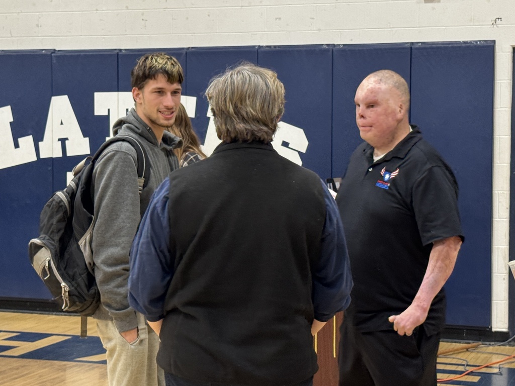 A man with visible burn scars, wearing a black polo shirt, speaks with two people—including a student with a backpack—inside the gym.