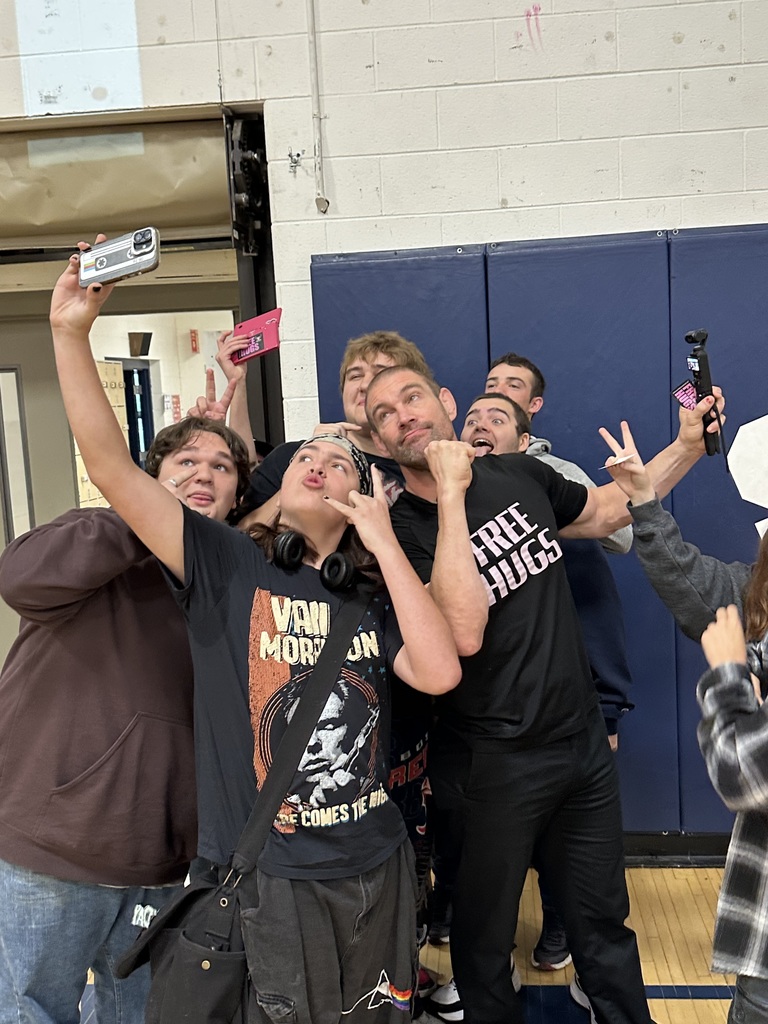 A group of students takes a selfie with the “Free Hugs” speaker in the gym. Everyone is laughing and making playful expressions while holding up their phones.
