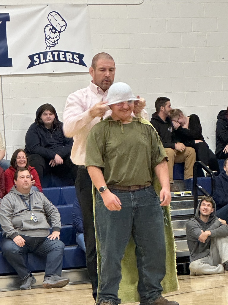 The speaker, wearing a pink dress shirt, places a colander-style helmet on a smiling student’s head during a gym presentation. Audience members in the bleachers laugh and watch.