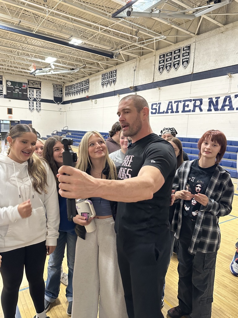 The “Free Hugs” presenter stands with a group of smiling students in the gym, holding up a camera for a selfie. The words “Slater Nation” are visible on the wall behind them.