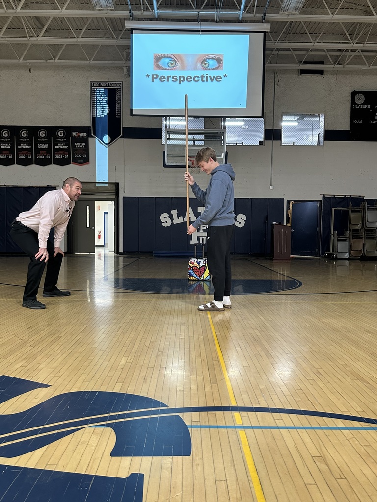 A presenter crouches in front of a student holding a tall stick on the gym floor, with the word “Perspective” projected on a screen behind them.