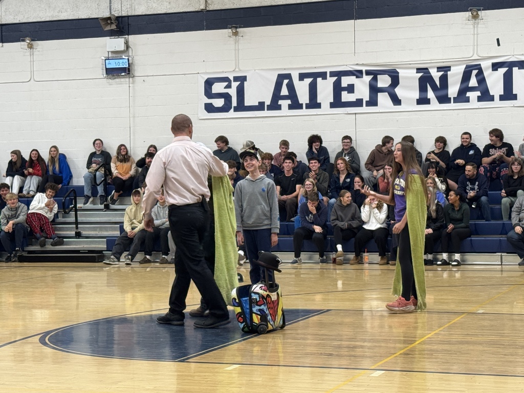 The presenter interacts with two students wearing green capes in the gym as a crowd of students watches and smiles from the bleachers.