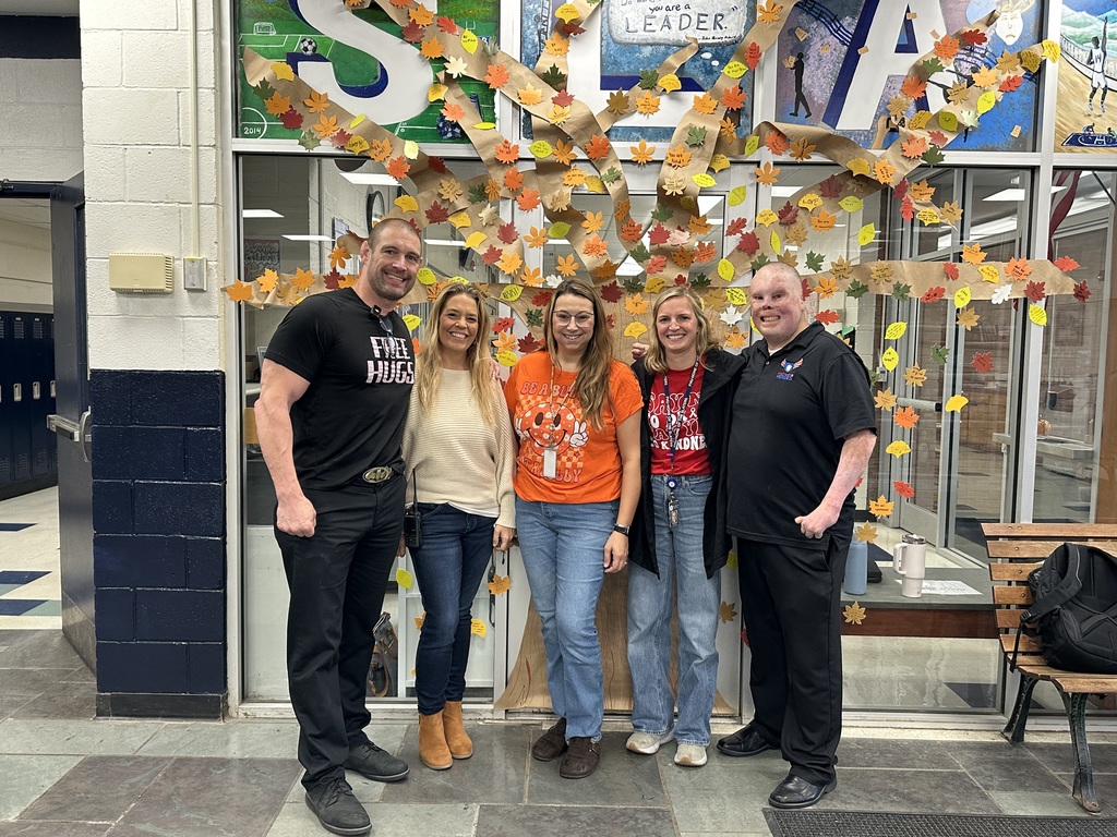 Five adults standing in front of a paper “kindness tree” decorated with colorful leaves. Two people wear orange shirts with kindness messages, and others wear neutral or black tops. The backdrop displays large letters spelling “SLATE.”