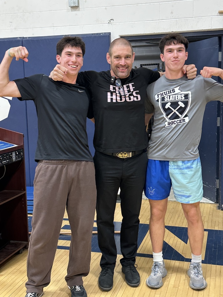 A man wearing a “Free Hugs” shirt stands in the gym with two students on either side, all smiling and flexing their arms for a photo. One student wears a black shirt, the other a Fair Haven Slaters soccer shirt.