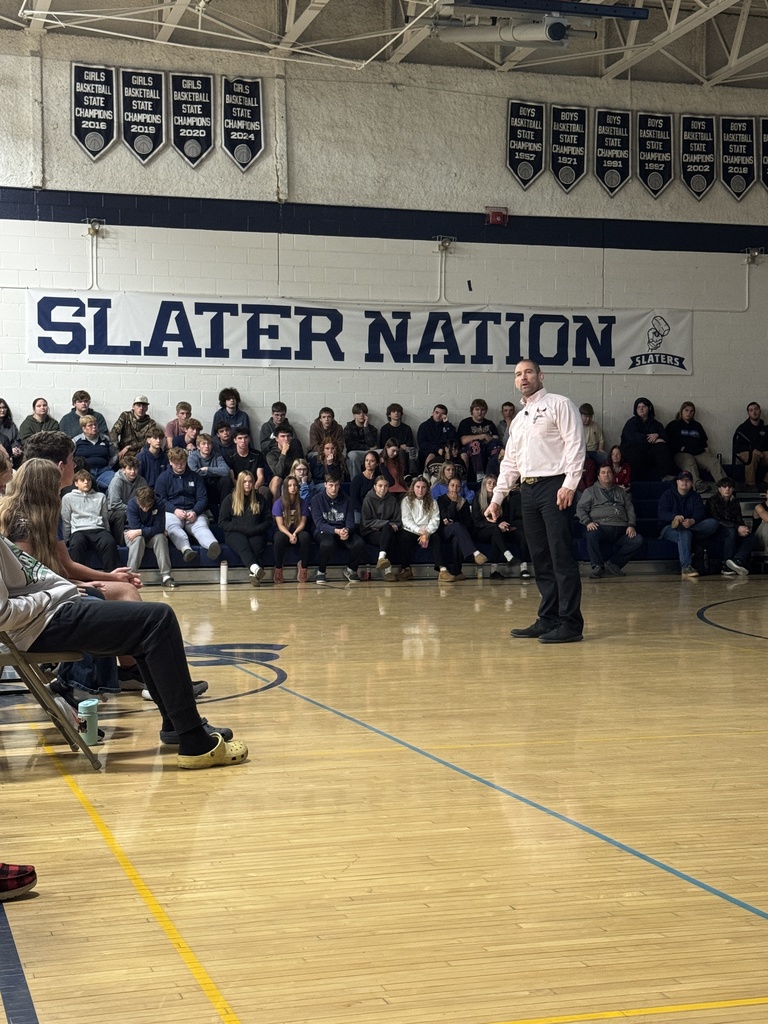 The speaker stands mid-presentation on the gym floor, addressing students seated in the bleachers under a “Slater Nation” banner.