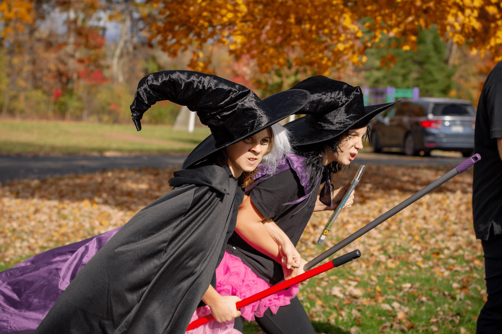 Two children dressed as witches in black hats and capes pretend to fly on broomsticks during the parade. Autumn leaves fill the background under bright sunlight.