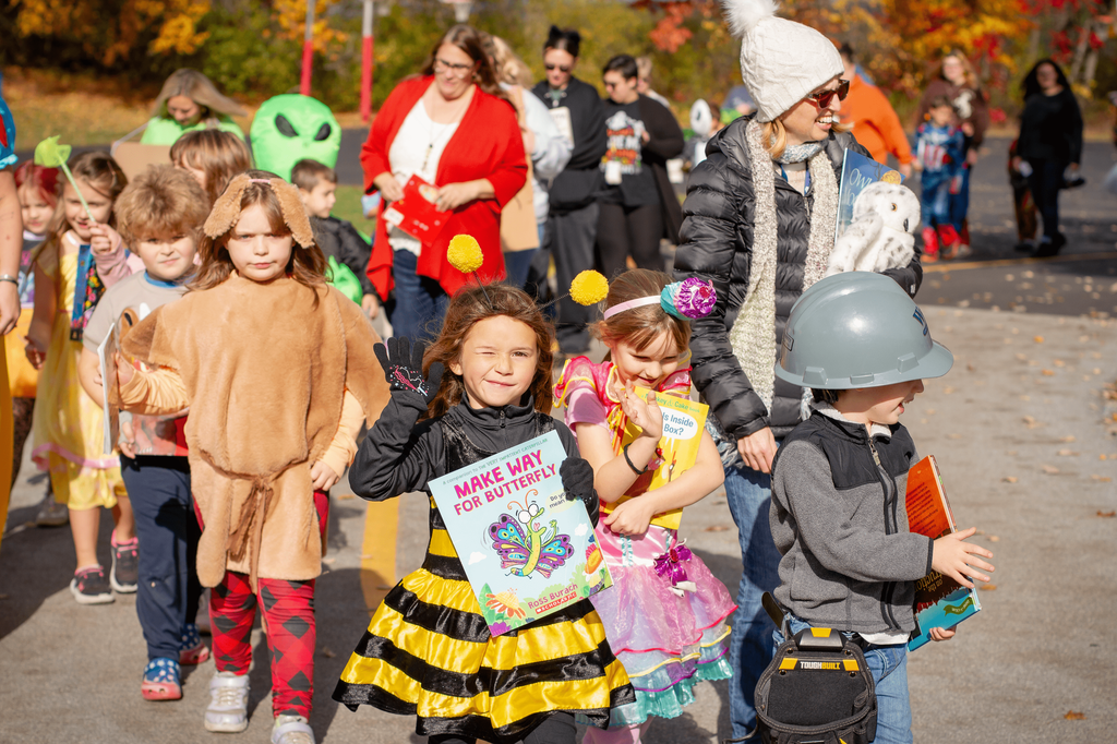 A group of young students parade outdoors in colorful costumes. One girl dressed as a bee waves while holding Make Way for Butterfly, followed by classmates as adults watch and guide them.