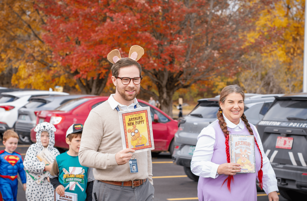Two adults dressed as Arthur characters hold Arthur’s New Puppy and Arthur Writes a Story. They smile and walk with children dressed as superheroes and storybook characters.
