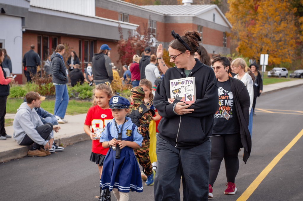 A woman wearing cat ears and glasses walks with children in costume, holding the book “Bad Kitty: Tasty Treats.” Kids are dressed as a police officer, a football player, and other characters, parading past families.