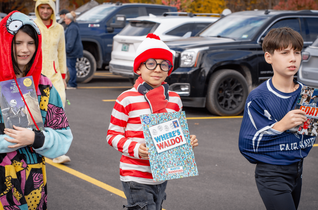 Three children in costumes walk in the parade. One dressed as “Where’s Waldo” holds the matching book, while others hold The Nightmare Before Christmas and Baseball GOAT.