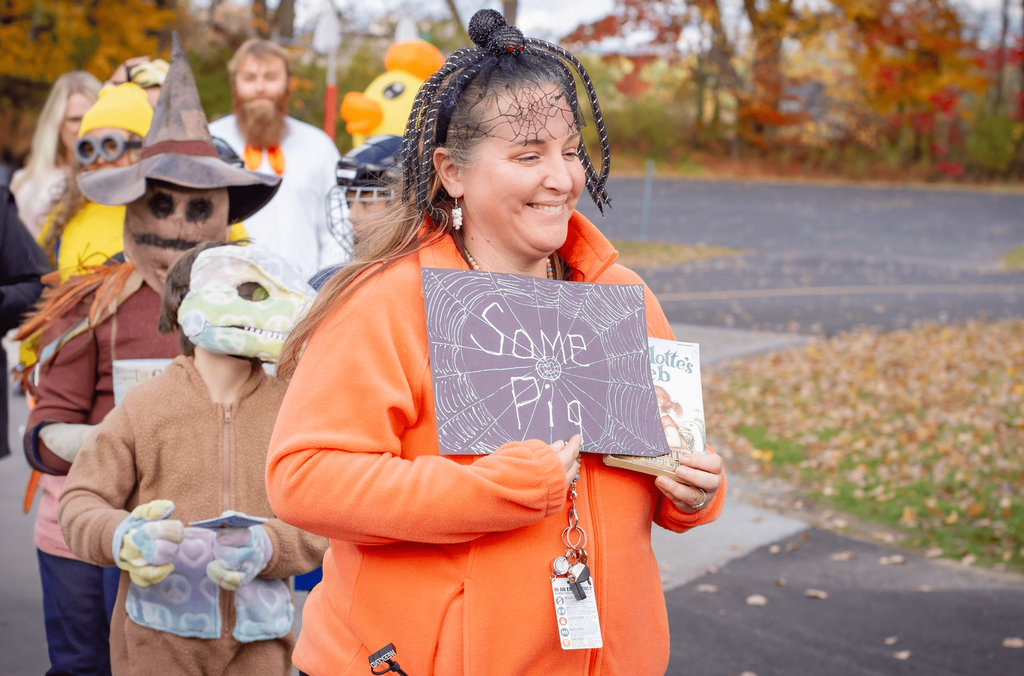 A smiling woman dressed in orange holds a “Charlotte’s Web” book and a handmade sign reading “Some Pig.” She wears a spider headband, walking in a parade with children in costumes behind her.
