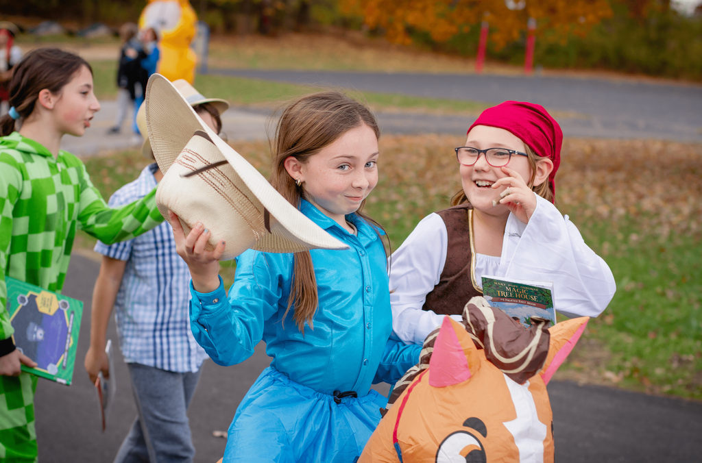 Two girls smile and pose during the parade — one dressed as a cowgirl tipping her hat, and the other as a pirate holding a Magic Tree House book. Other costumed students walk behind them.