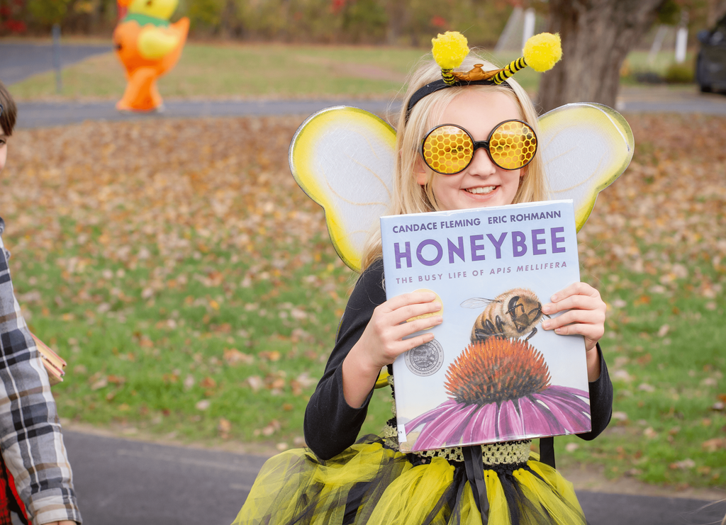 A young girl dressed as a bee smiles while holding the book Honeybee: The Busy Life of Apis Mellifera. She wears a yellow-and-black costume with wings and large honeycomb-patterned glasses.