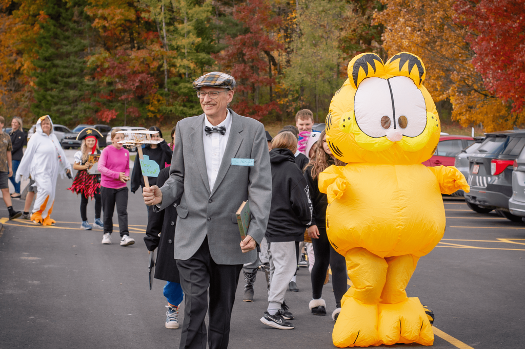 A man dressed as author Orville Wright holds a prop sign reading “Fly, fly, fly!” while walking beside a large inflatable Garfield costume. Other participants in creative outfits follow behind.
