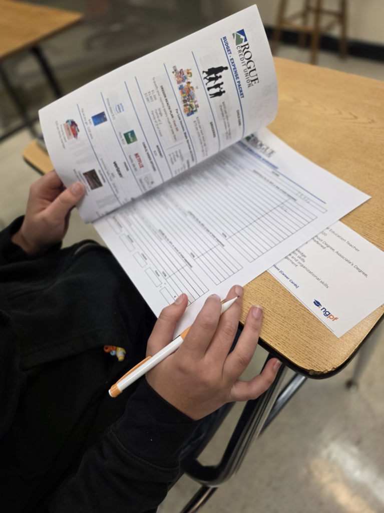 A student holds a pencil and reviews a printed packet titled “Rogue Credit Union Budget Expense Project” while seated at a classroom desk.