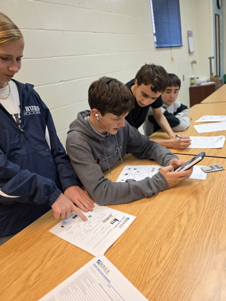 Four students collaborate at a classroom table, using calculators and worksheets for a financial literacy activity. One student points at the paper while another checks figures on a phone.