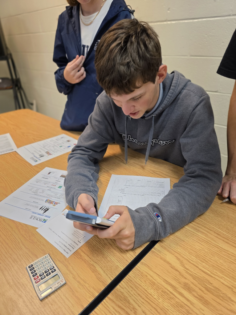 A student in a gray hoodie uses a phone calculator to fill out a budgeting worksheet as another student stands beside, observing the work.