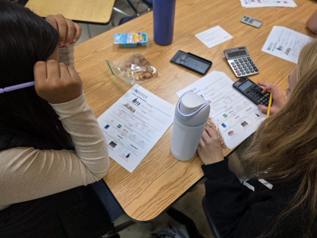 Two students seated at a desk use calculators and fill in a “Rogue Credit Union” worksheet. A water bottle and snack bag are on the table.
