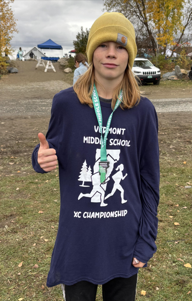 A student wearing a yellow knit hat and a navy “Vermont Middle School XC Championship” shirt smiles and gives a thumbs-up while standing outdoors with a race medal around their neck.