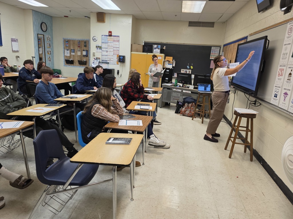 A teacher explains a concept at the smartboard while students listen attentively in a classroom setting.