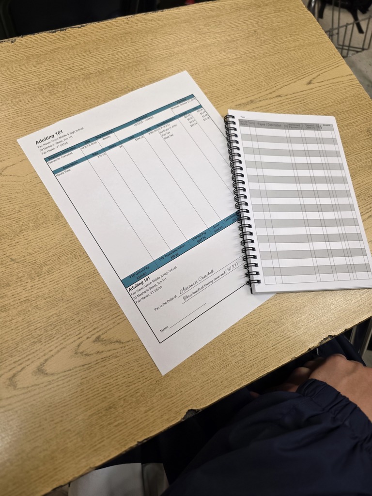Close-up of a student’s desk showing an “Adulting 101” worksheet and checkbook register for a budgeting exercise.