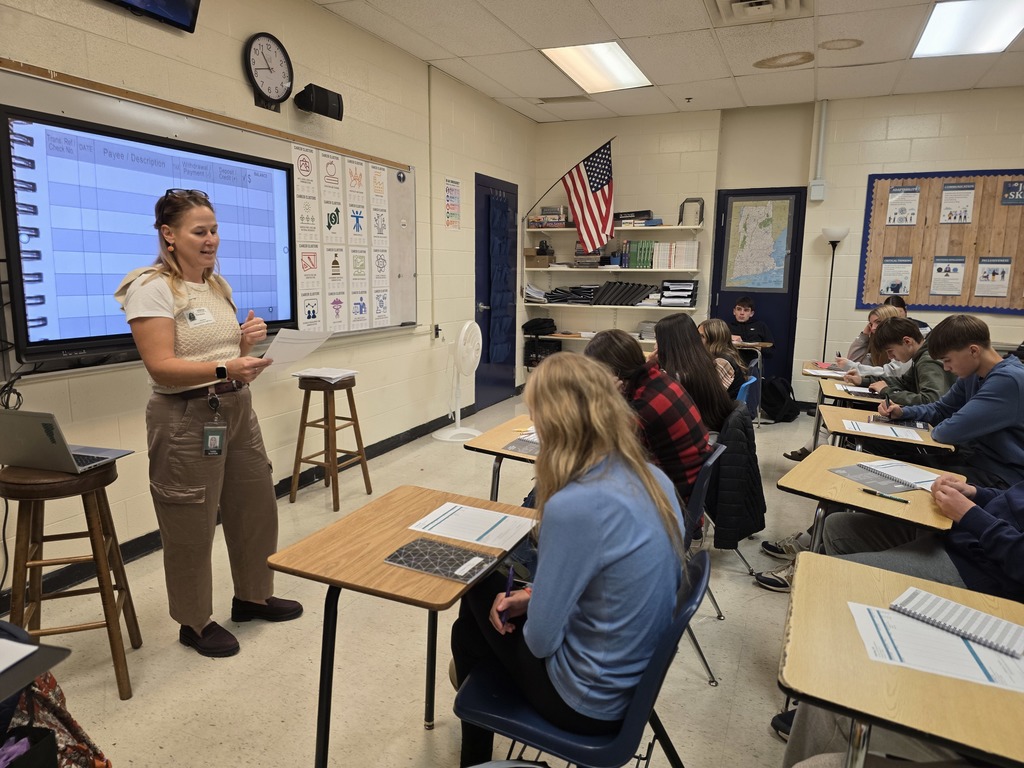 The teacher stands at the front of the room speaking to students as they take notes in an “Adulting 101” class.