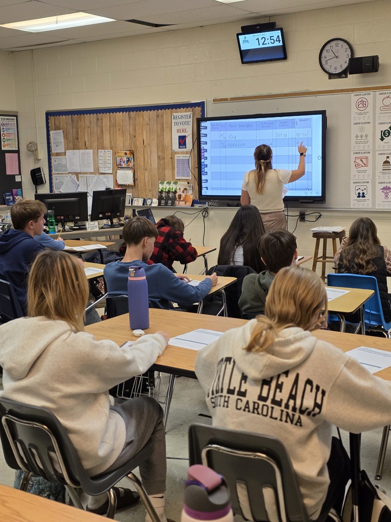Students sit at desks facing a teacher who writes on a digital smartboard during a classroom lesson.