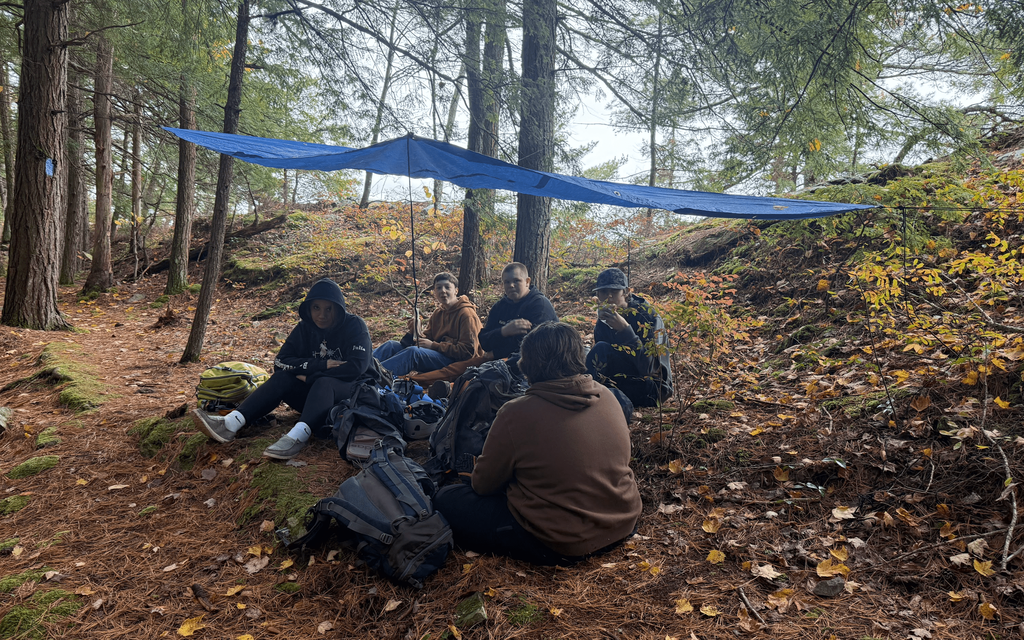 A group of students rests under a tarp shelter in the woods during a fall outdoor trip.