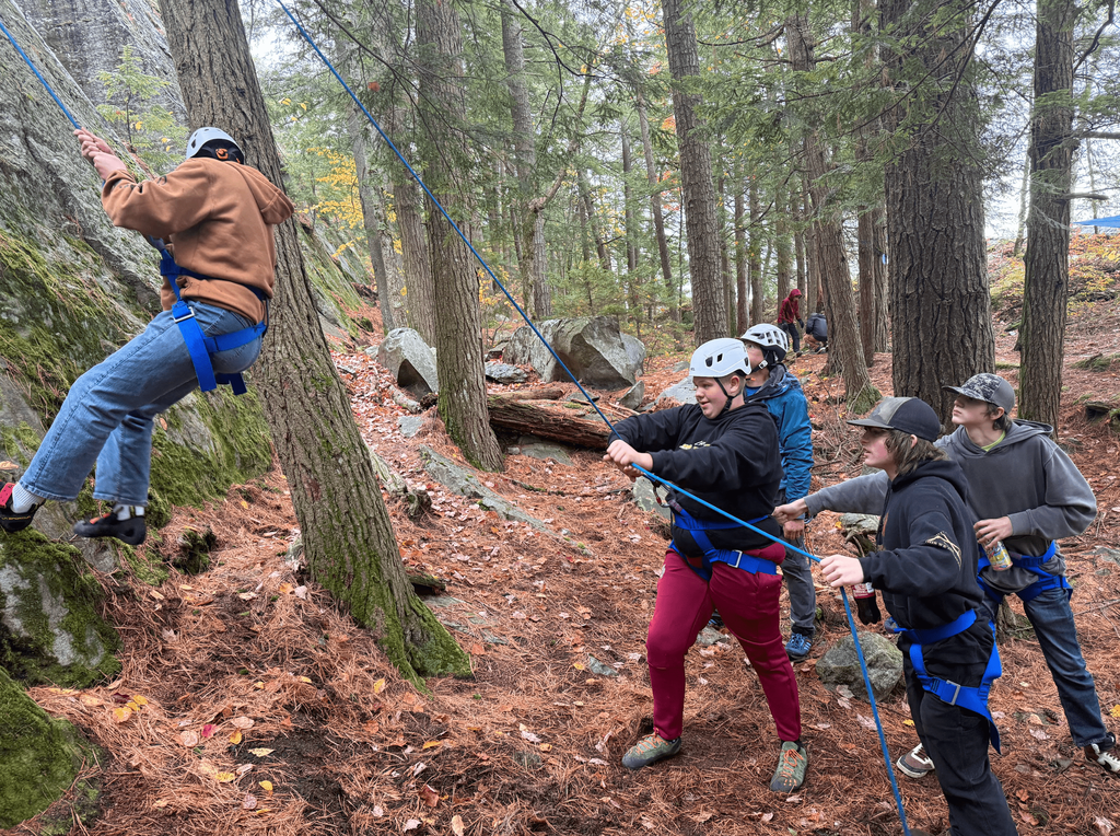 Students wearing helmets and harnesses work together belaying a classmate climbing a rock wall in a forest.