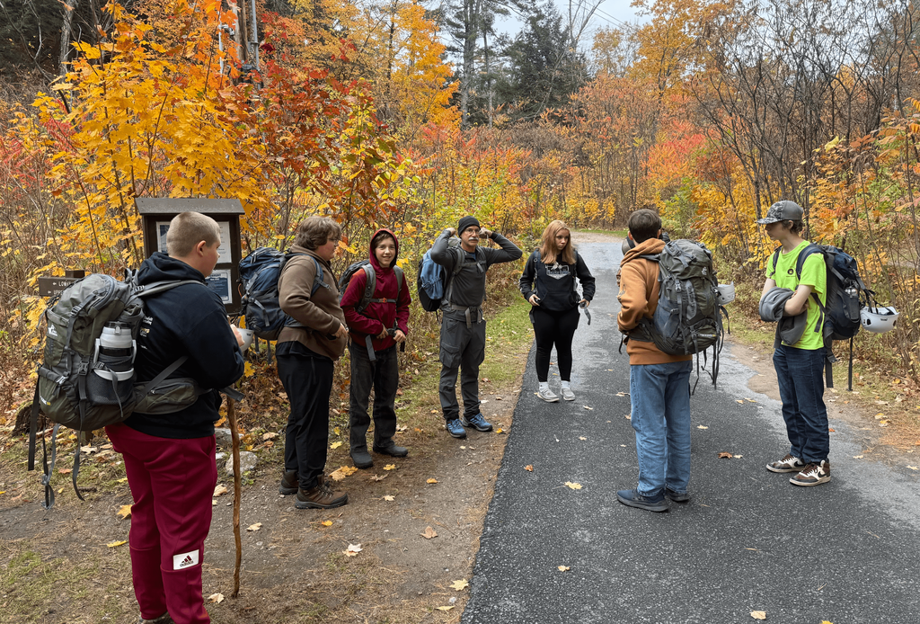 A group of students with backpacks gathers at a trailhead surrounded by vibrant fall foliage.