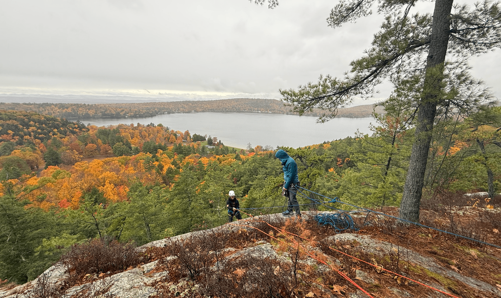 Two students with ropes and helmets stand atop a rocky cliff overlooking a lake and colorful autumn trees.