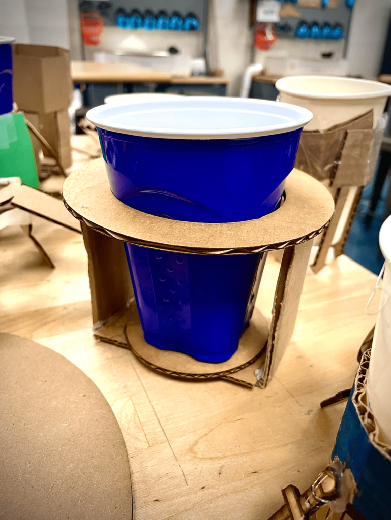 A close-up of a blue plastic cup supported by a cardboard holder created as part of a student design project.