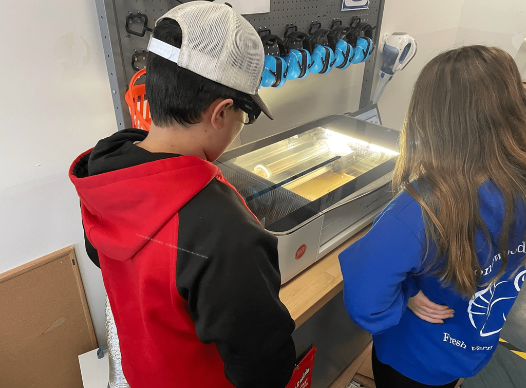 Two students wearing safety glasses observe a laser engraver in operation inside a school makerspace.
