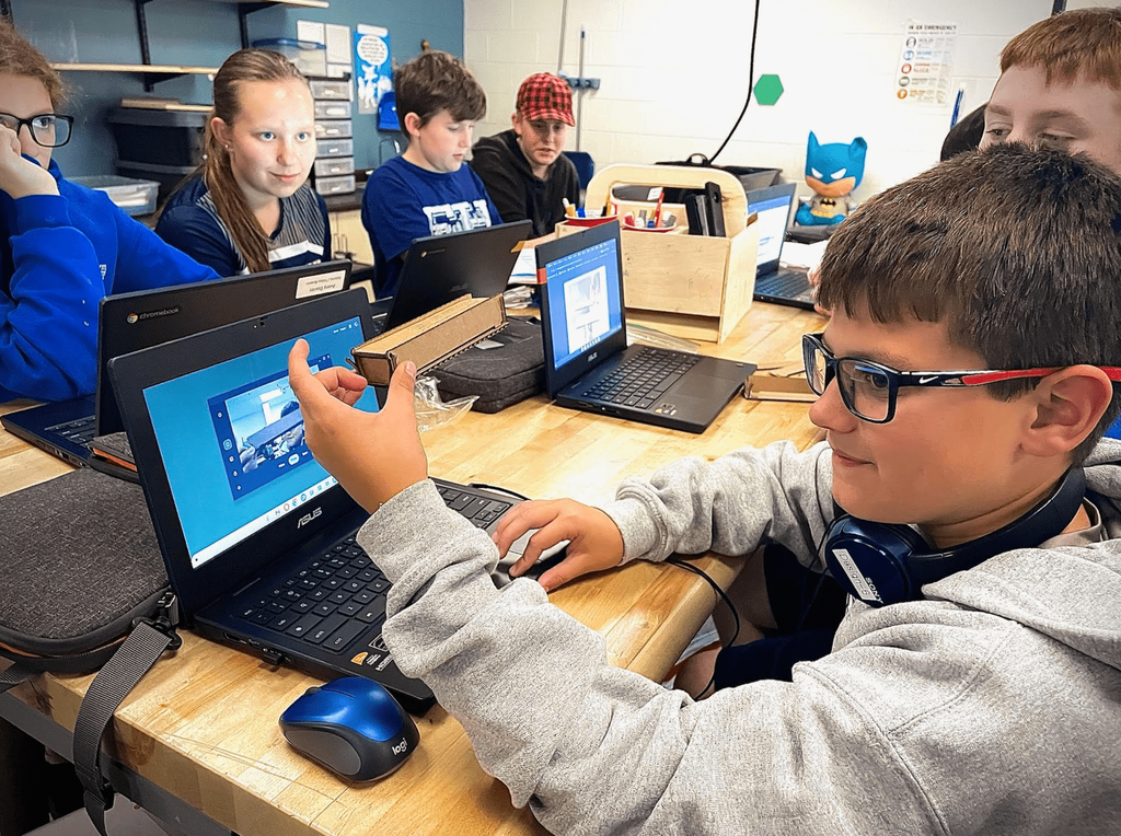 Students sit at a table using laptops and examining small cardboard pieces during a hands-on design activity.