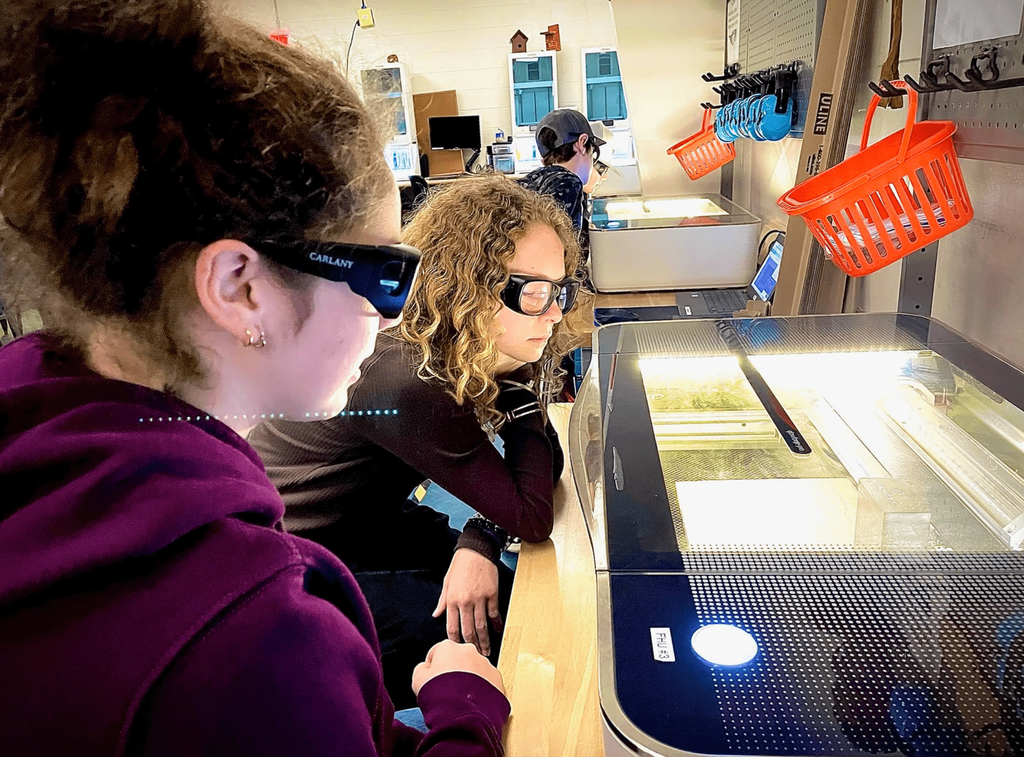 Two students wearing protective glasses watch a laser engraver as it cuts material for a design project.