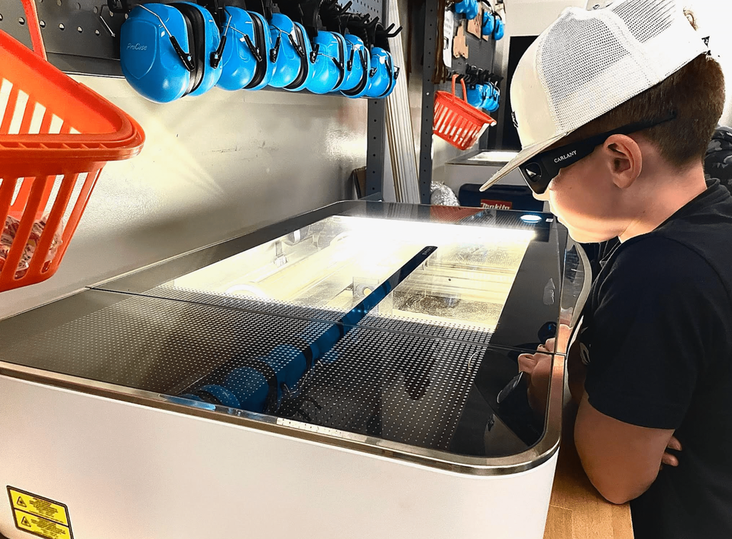A student wearing a white hat and safety glasses closely watches a Glowforge laser engraver in use.