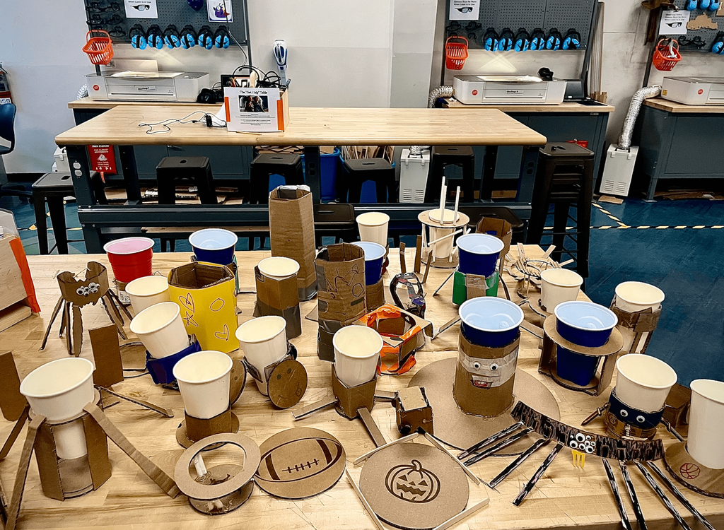 A table displays multiple student-made cardboard and cup creations, including pumpkins, spiders, and other imaginative designs, in a school makerspace.
